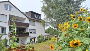 A residential building with a balcony, a garden with sunflowers, and a wooden play structure. - Large cozy apartment "Ziegenpeter" (DG = 2nd floor) - Kolvenbacher Ziegenhof 95sqm (Hohn)