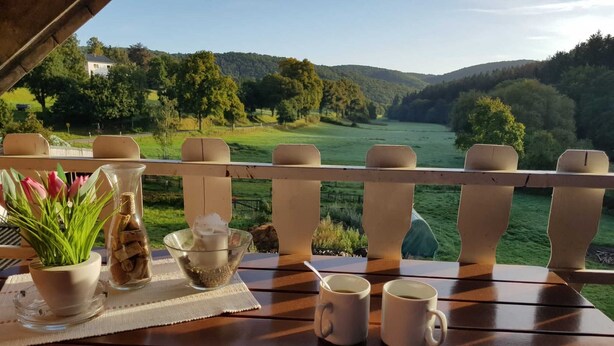 A wooden deck with a table set for two, overlooking a scenic landscape with hills and trees. - Large cozy apartment "Ziegenpeter" (DG = 2nd floor) - Kolvenbacher Ziegenhof 95sqm (Hohn)