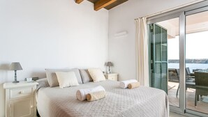 Quarto com uma cama grande, duas mesas de cabeceira com luminárias, vista para o mar através de uma porta de vidro deslizante e uma varanda com mobiliário exterior.