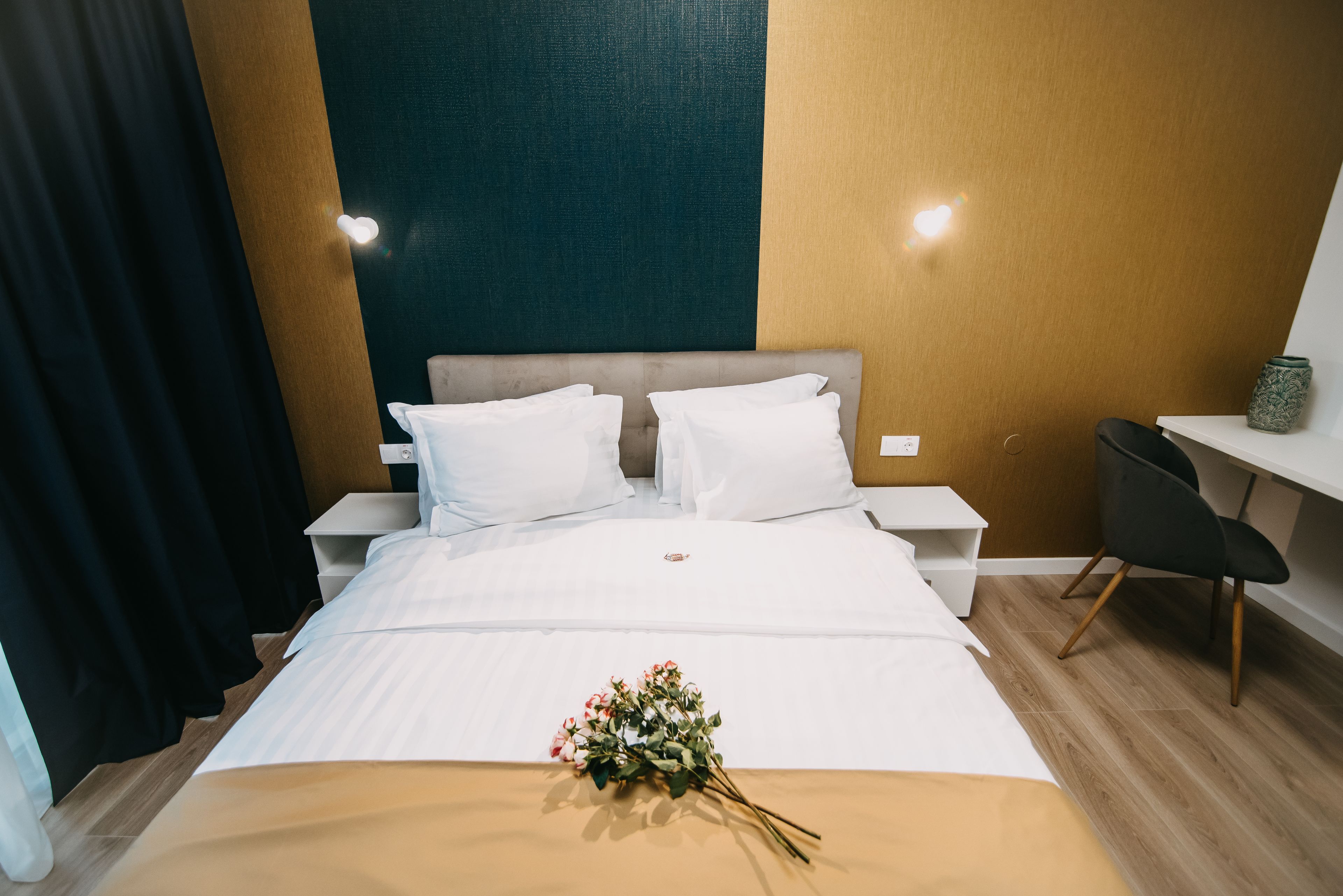 A neatly made bed with white linens and a bouquet of flowers, a nightstand with a lamp, a desk with a chair, and a wall with a textured pattern.