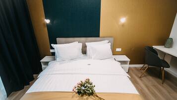 A neatly made bed with white linens and a bouquet of flowers, a nightstand with a lamp, a desk with a chair, and a wall with a textured pattern.