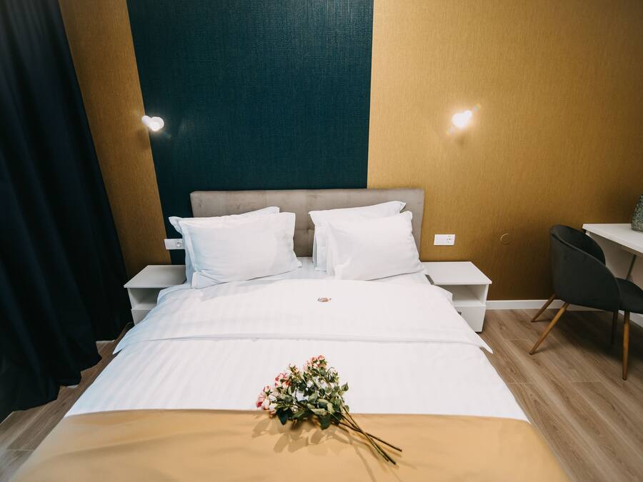 A neatly made bed with white linens and a bouquet of flowers, a nightstand with a lamp, a desk with a chair, and a wall with a textured pattern.
