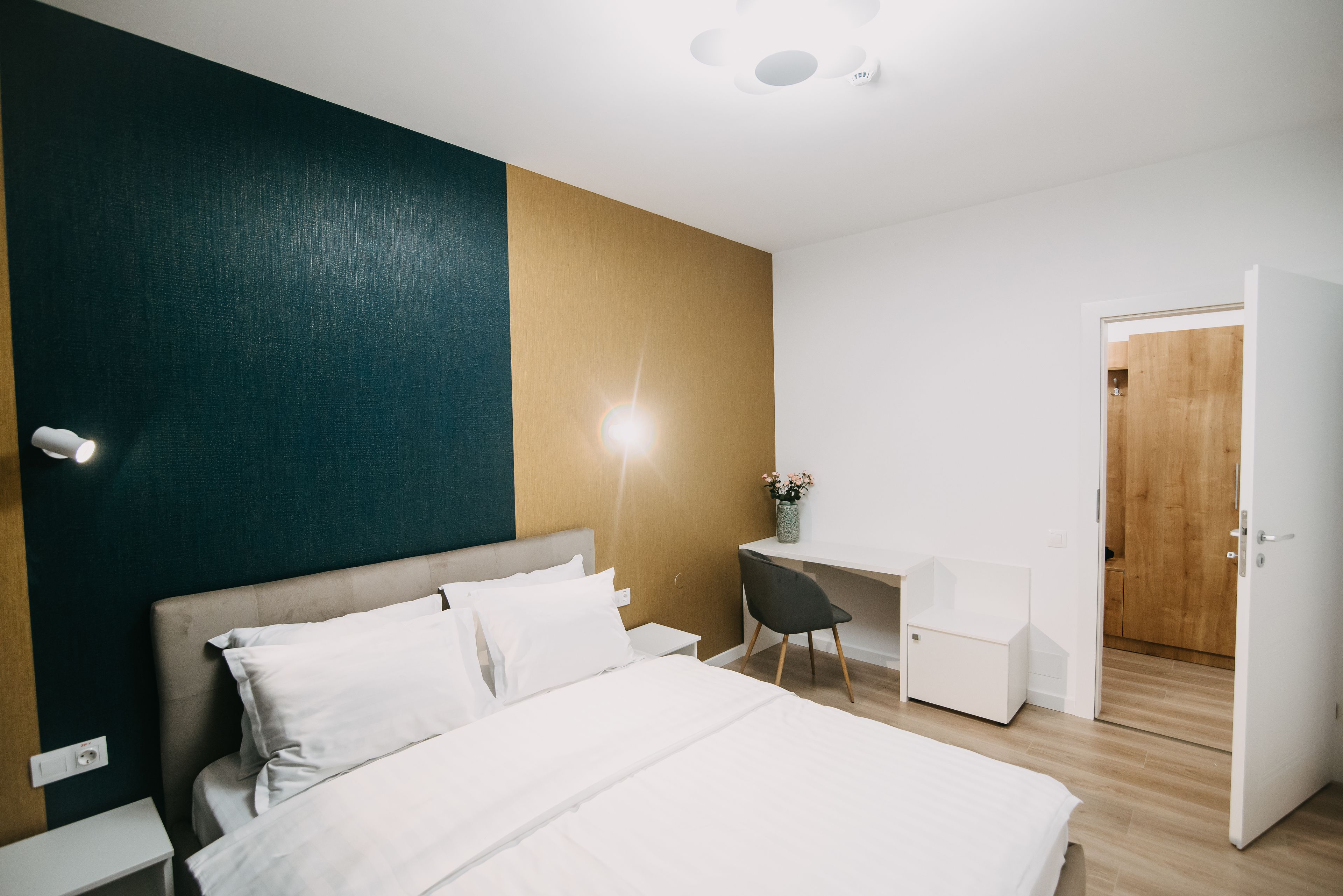 A modern bedroom with a bed, bedside table, desk, chair, and a door leading to another room.