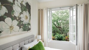 Un dormitorio con un ventanal grande, una cama con ropa de cama blanca, un cabecero tapizado en gris y un cuadro floral en la pared.