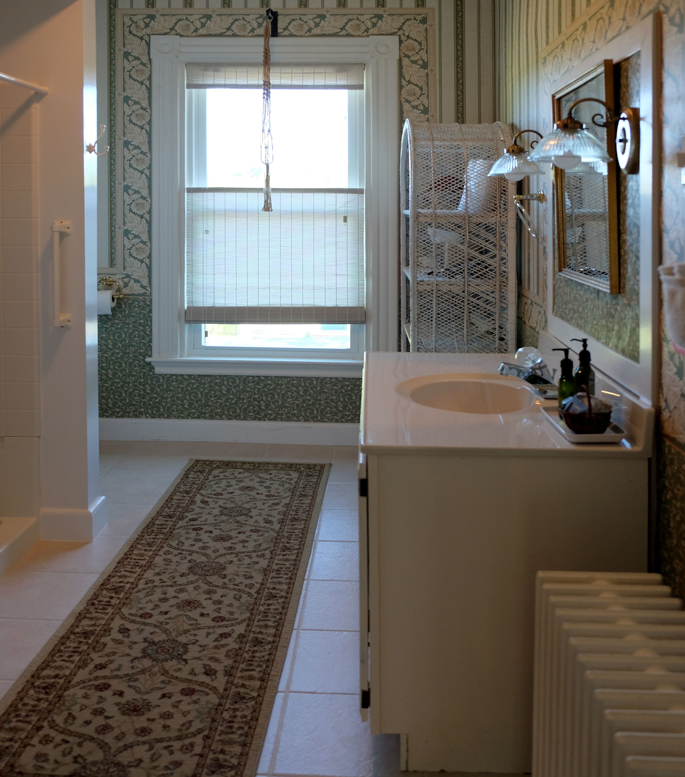 Un baño con un lavamanos blanco, un espejo, una ventana con persianas y una alfombra estampada.