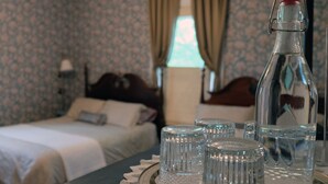A hotel room with a neatly made bed, a bedside table with a tray holding glasses and a bottle, and a window with curtains.