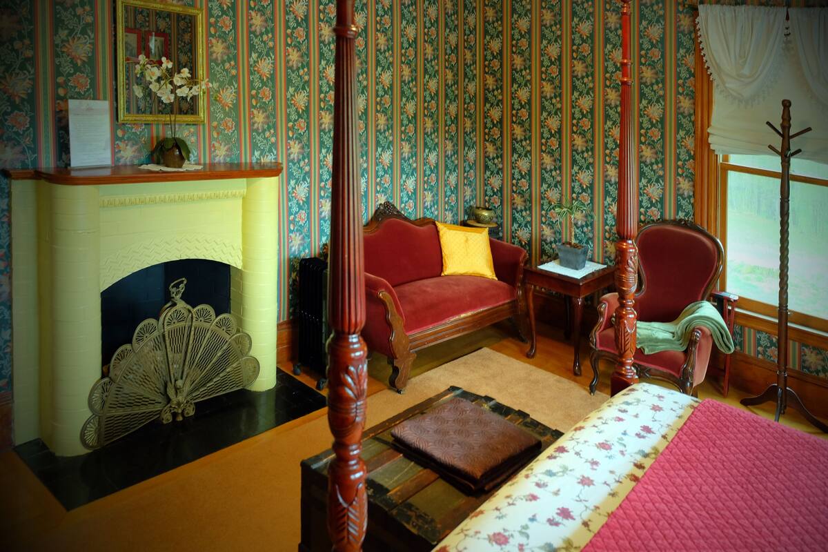 A room with a four-poster bed, a red sofa, a fireplace, and floral wallpaper.