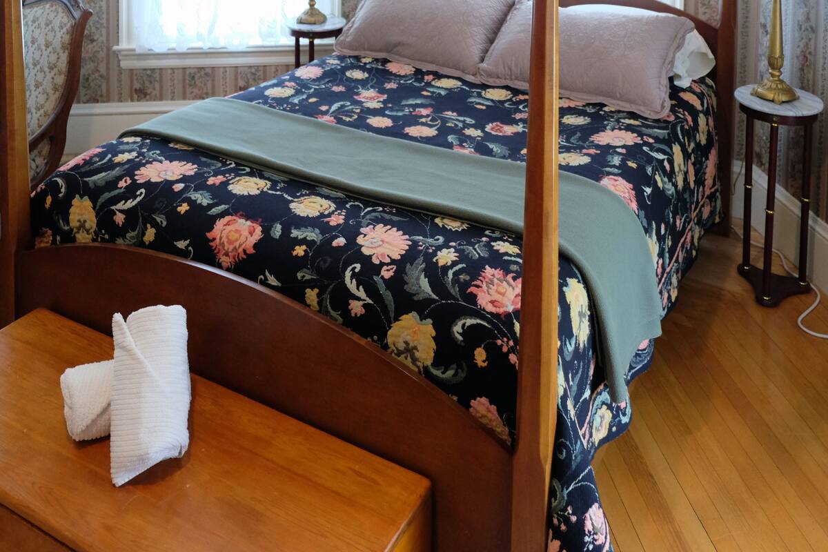 A four-poster bed with floral bedding, a wooden bench with a towel, and a bedside table with a lamp.