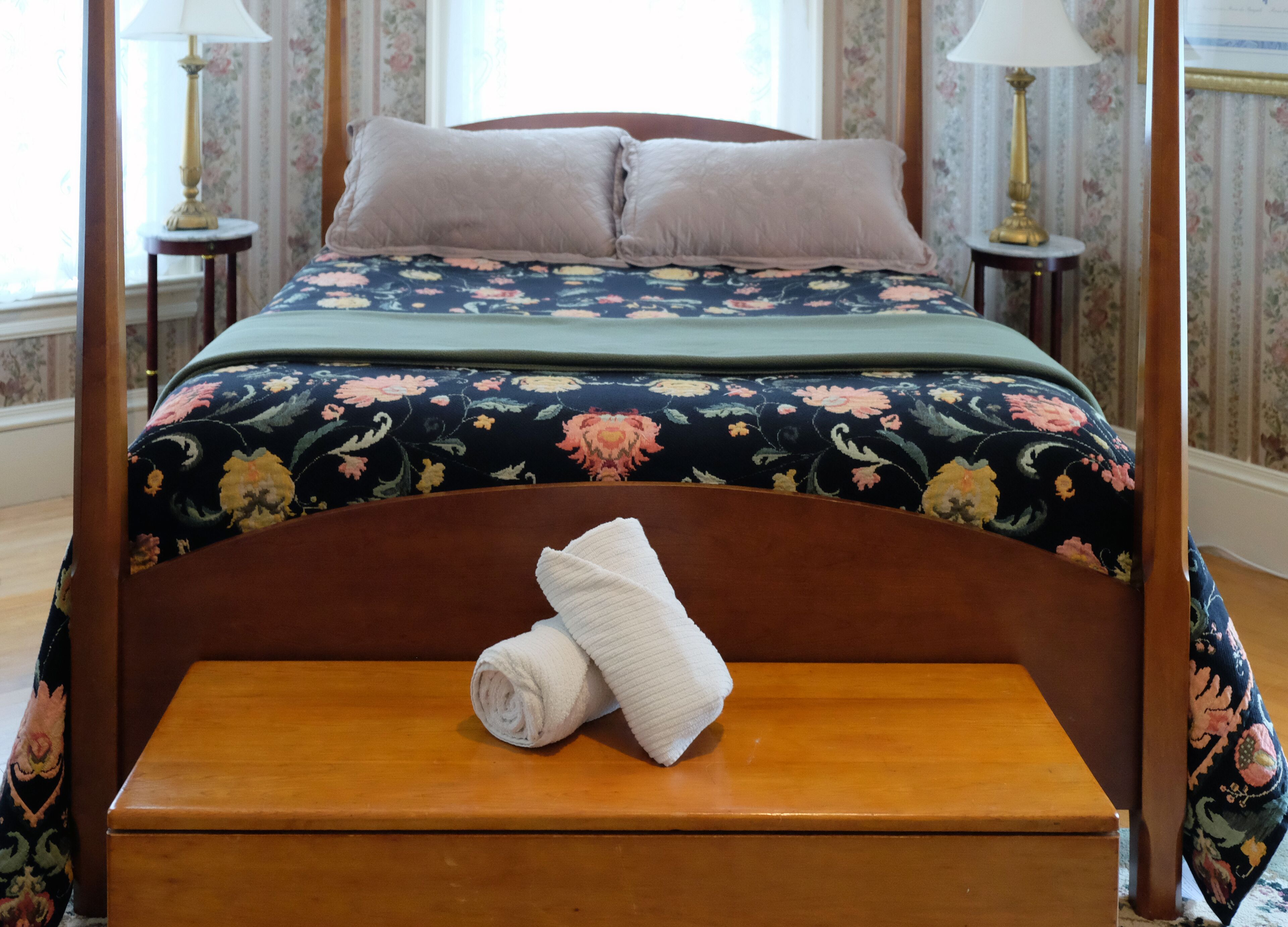A four-poster bed with floral bedding, two pillows, and a rolled towel on a wooden bench.