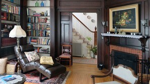 A traditional living room with a fireplace, bookshelves, a leather armchair, and a staircase leading to another room.