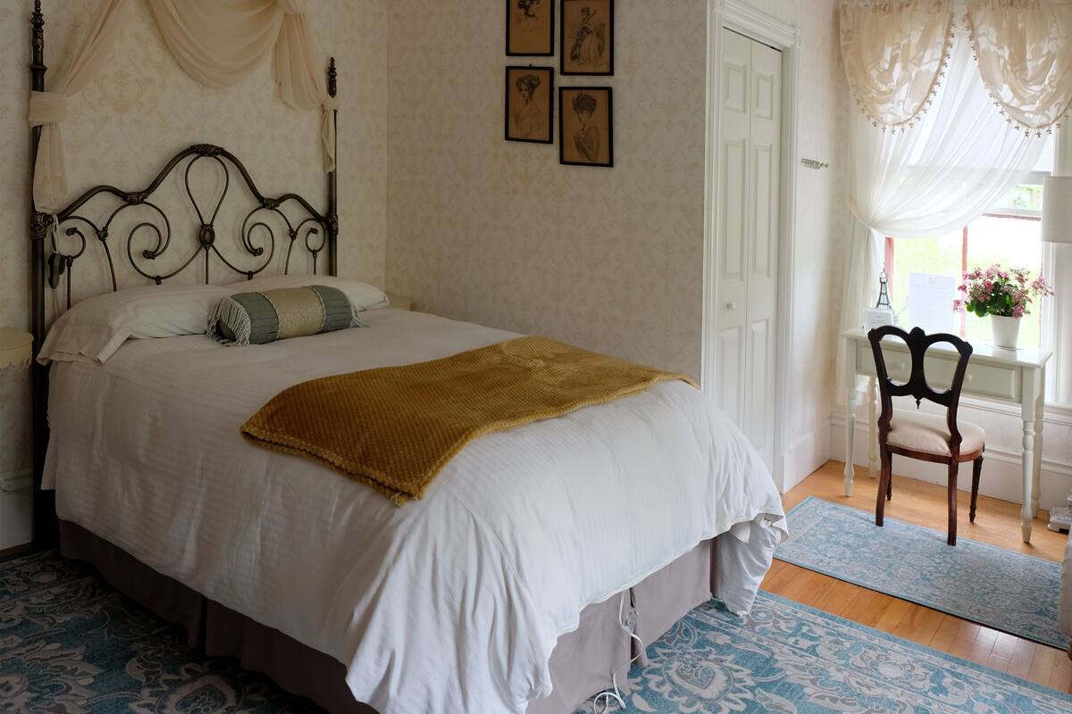 A neatly made bed with a white and brown blanket, a wooden headboard, and a bedside table with a lamp. There are framed pictures on the wall and a window with sheer curtains.