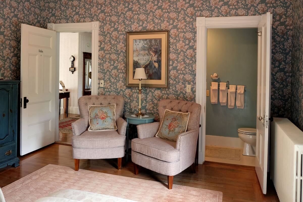 A room with two armchairs, a small table, a framed picture, a radiator, and a door leading to a bathroom.