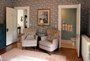 Une pièce comprenant deux fauteuils, une petite table, un tableau encadré, un radiateur et une porte donnant sur une salle de bain.