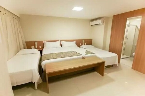 A hotel room with a double bed, a single bed, a wooden bench, and an air conditioning unit.