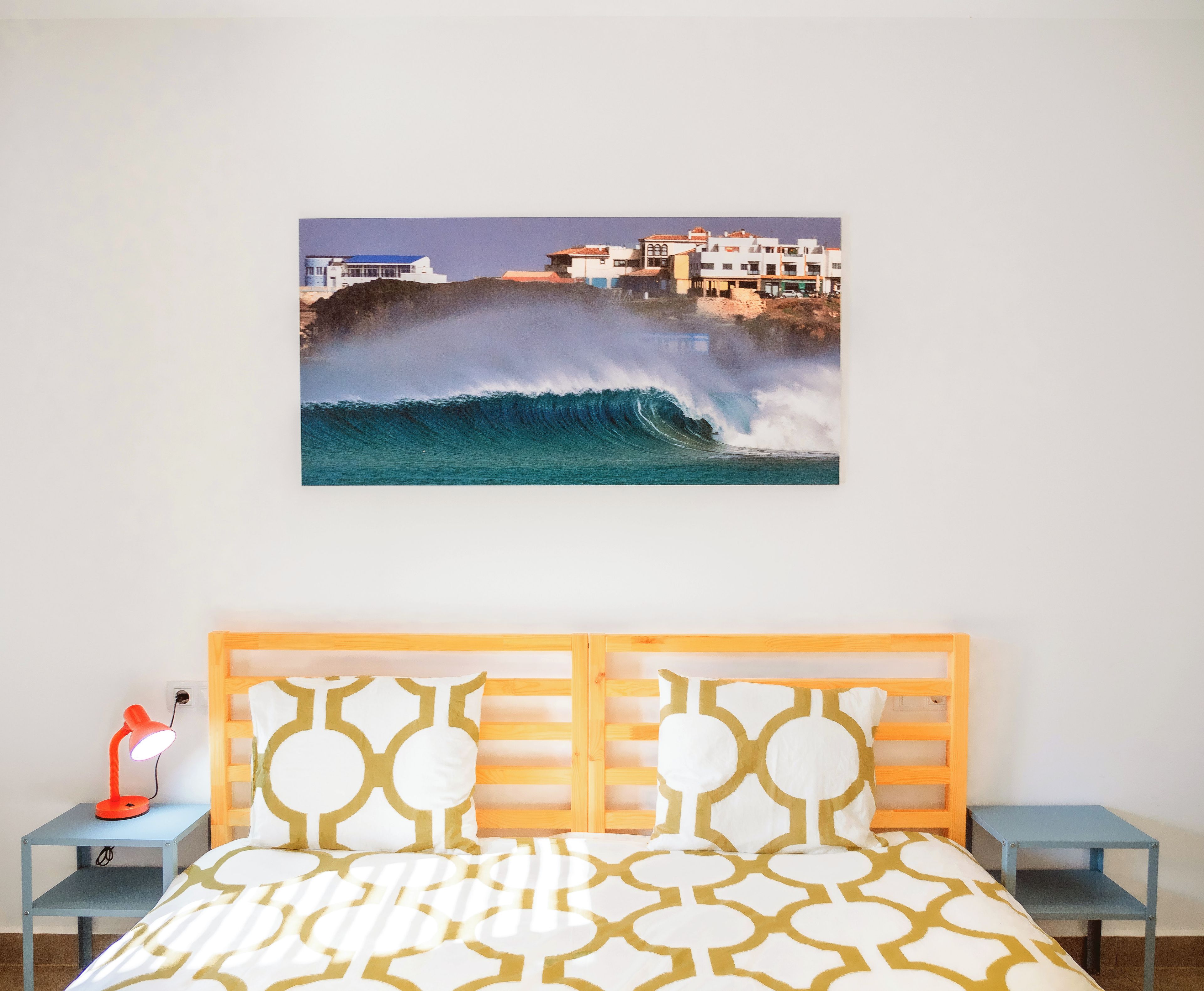 Une chambre à coucher comprenant un lit, une table de chevet, une lampe et un tableau représentant un paysage côtier, accroché au mur.
