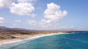 A coastal landscape with a beach, clear blue sea, and distant mountains under a partly cloudy sky. - MAAR House (La Oliva)