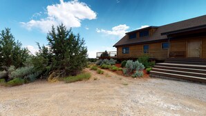 A wooden cabin with a gravel driveway, surrounded by evergreen trees and shrubs. - Mustang Mesa Amazing Views (Blanding)