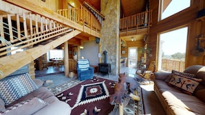 A spacious log cabin interior with a high ceiling, a stone fireplace, a wooden staircase, and a balcony with a view. - Juniper Ridge Family Cabin (Blanding)