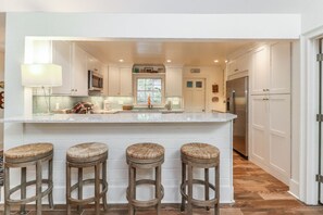 A kitchen with white cabinets, a large island with stools, a microwave, and a refrigerator. - Beautifully Updated Three Bedroom Sea Pines Home ~ Short Walk to Beach (Hilton Head Island)