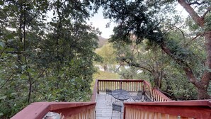 A wooden deck with metal chairs and a table, surrounded by dense foliage and a view of a lake. - The Cabin at Lydias Canyon (Glendale)