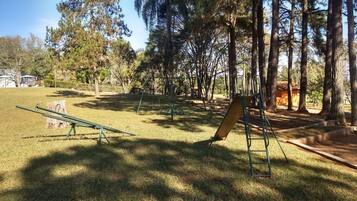 Explore the serene park area with swings and a seesaw surrounded by tall trees and a lush green field.