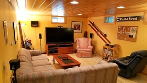 Ein gemütliches Wohnzimmer mit einem großen Flachbildfernseher, einem rosa Sessel, einer Couch und einem Liegesessel. Ski lehnen an der Wand und ein Schwarzes Brett ist mit verschiedenen Gegenständen bestückt.