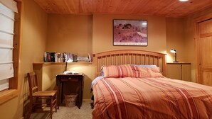 Ein gemütliches Schlafzimmer mit Holzbalkendecke, einem Bett mit gestreifter Bettwäsche, einem Nachttisch mit Lampe, einem Sessel und einem Bücherregal mit Büchern.