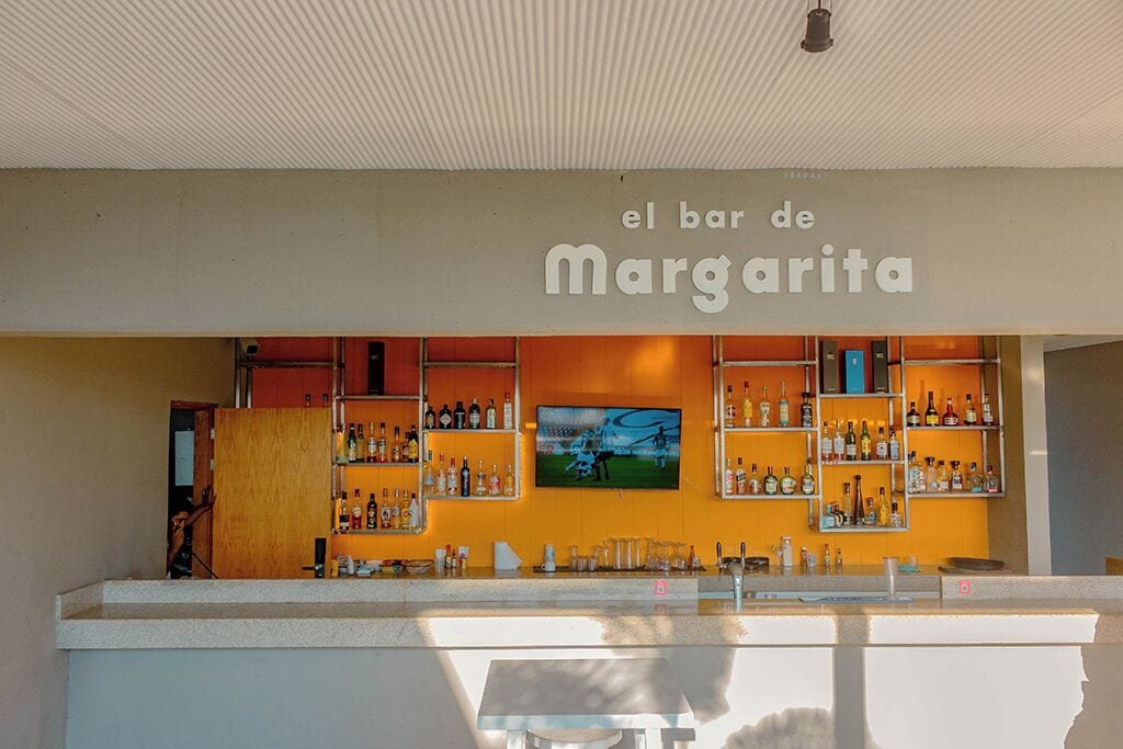 Un bar llamado 'el bar de Margarita' con estantes llenos de botellas y una pantalla de televisión.