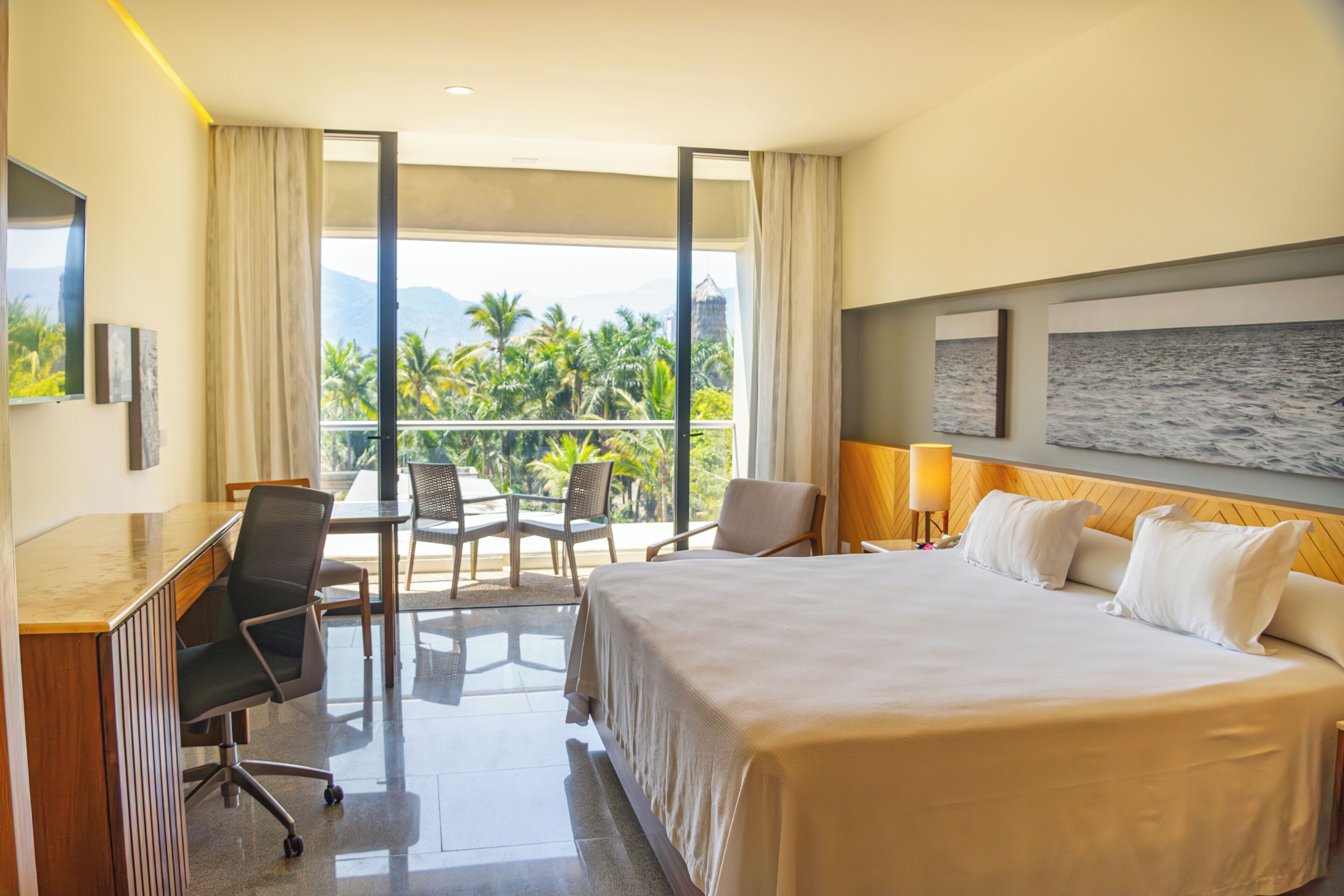 A hotel room with a bed, desk, chairs, a balcony with a view of palm trees and mountains.