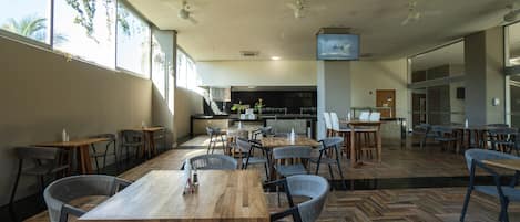 Un comedor amplio con varias mesas y sillas de madera, ventanas grandes, ventiladores de techo y una cocina al fondo.