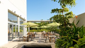 Un patio al aire libre con mesas y sillas, una pequeña fuente de agua y abundante vegetación.