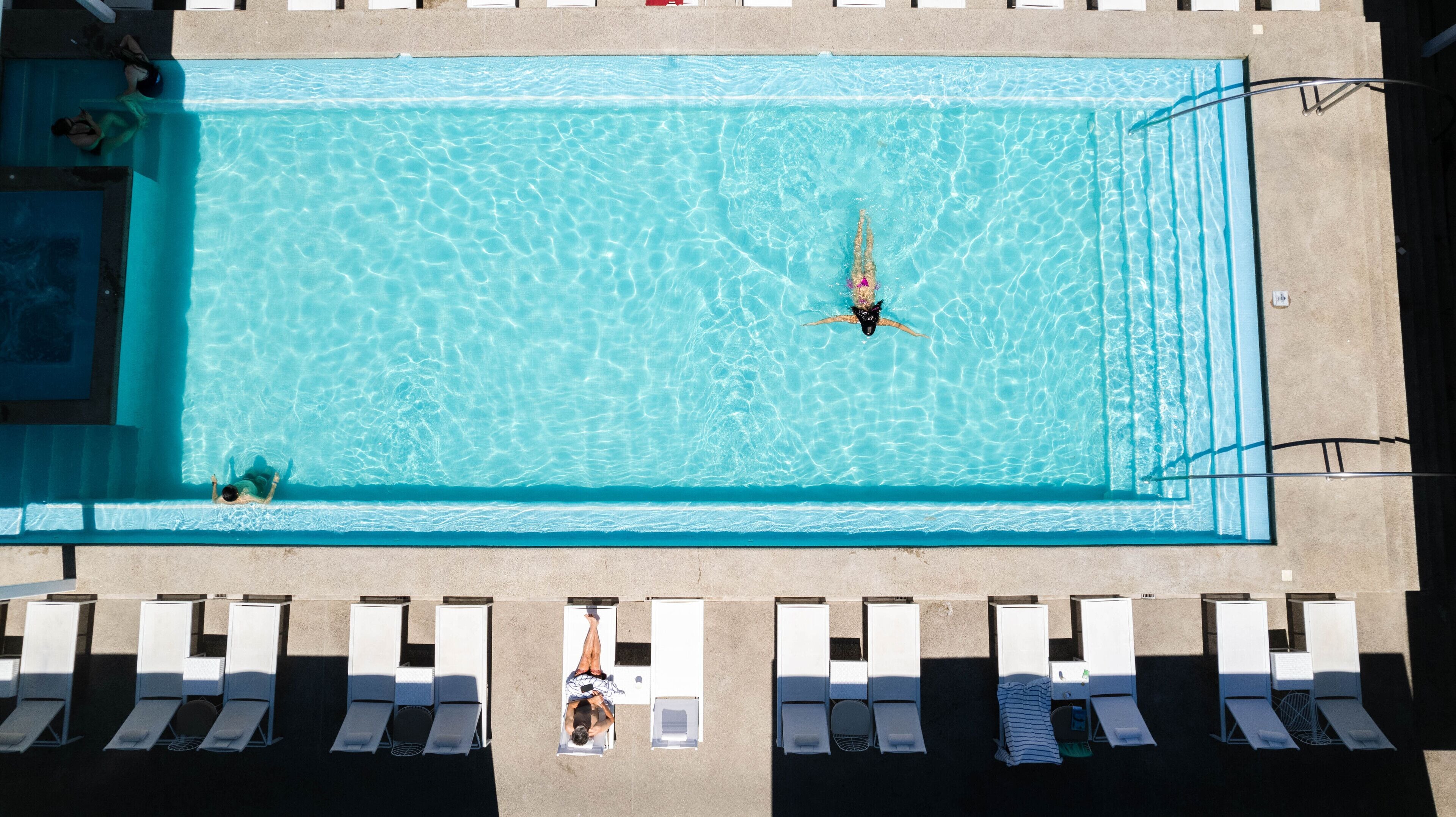 A rectangular swimming pool with clear blue water, surrounded by white sun loungers and a concrete deck.