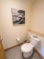 Un baño con inodoro, un cubo de basura, un rollo de papel higiénico y un cuadro de un paisaje marino rocoso en la pared.