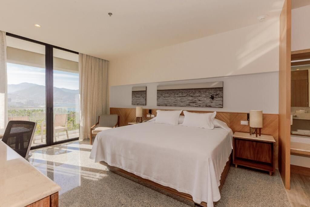 Habitación de hotel moderna con una cama grande, un escritorio, una silla y vistas a las montañas desde el balcón.