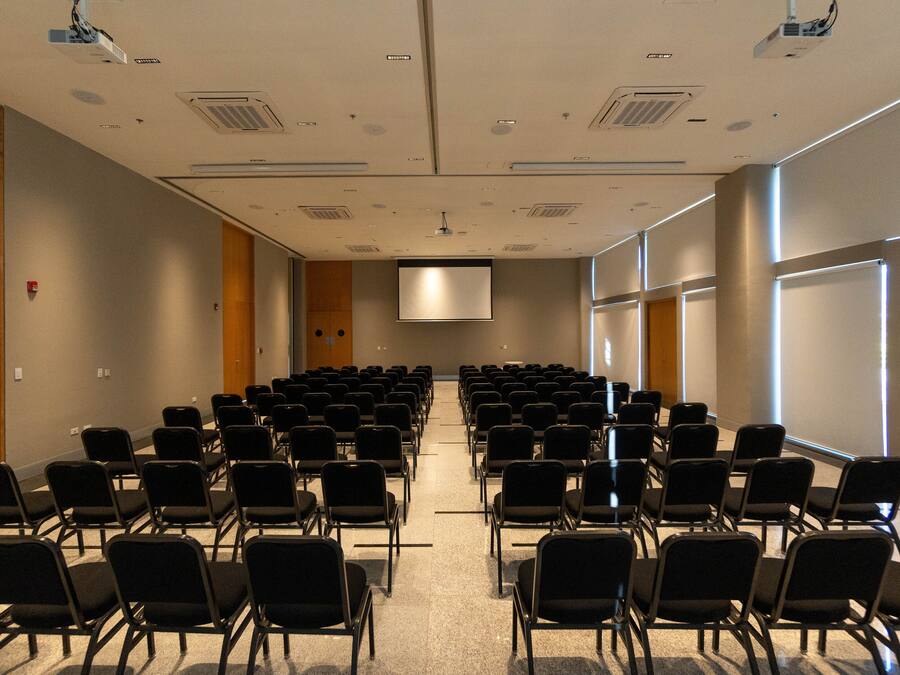 Una sala conferenze con file di sedie nere, uno schermo grande nella parte anteriore e finestre lungo una parete.