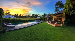 A pool with lounge chairs and umbrellas, a grassy lawn, and a building with outdoor seating. - Villa Artis (Keroboken Kelod)