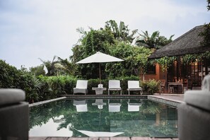 A pool area with lounge chairs and a table under a canopy, surrounded by lush greenery and a building with a thatched roof. - Villa Artis (Keroboken Kelod)