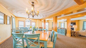 Un espace repas comprenant une grande table en bois et des chaises assorties, un lustre, et une vue sur l’extérieur grâce à de grandes fenêtres.