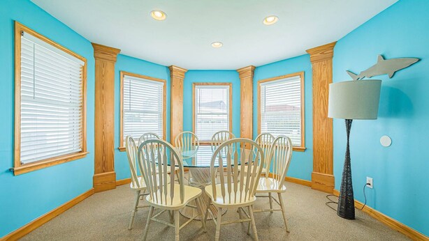 A dining room with a round table and six chairs, large windows with blinds, and a lamp on the right. - By the Sea - KDH MP 7.5, Oceanside, 320 Yds to Beach, Enclosed Pool, Hot Tub, Pets OK, Flex Stays (Kill Devil Hills)