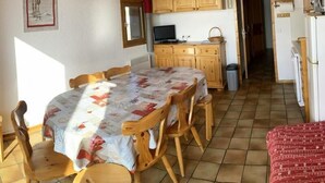 Un coin repas avec une table dressée pour quatre personnes, des chaises en bois, un téléviseur posé sur un meuble en bois, et un espace cuisine équipé d’un réfrigérateur et d’un four à micro-ondes.