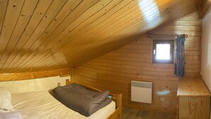 Une petite pièce chaleureuse avec un plafond en bois, un lit simple avec une literie blanche et grise, une petite fenêtre avec des rideaux et un radiateur blanc fixé au mur.