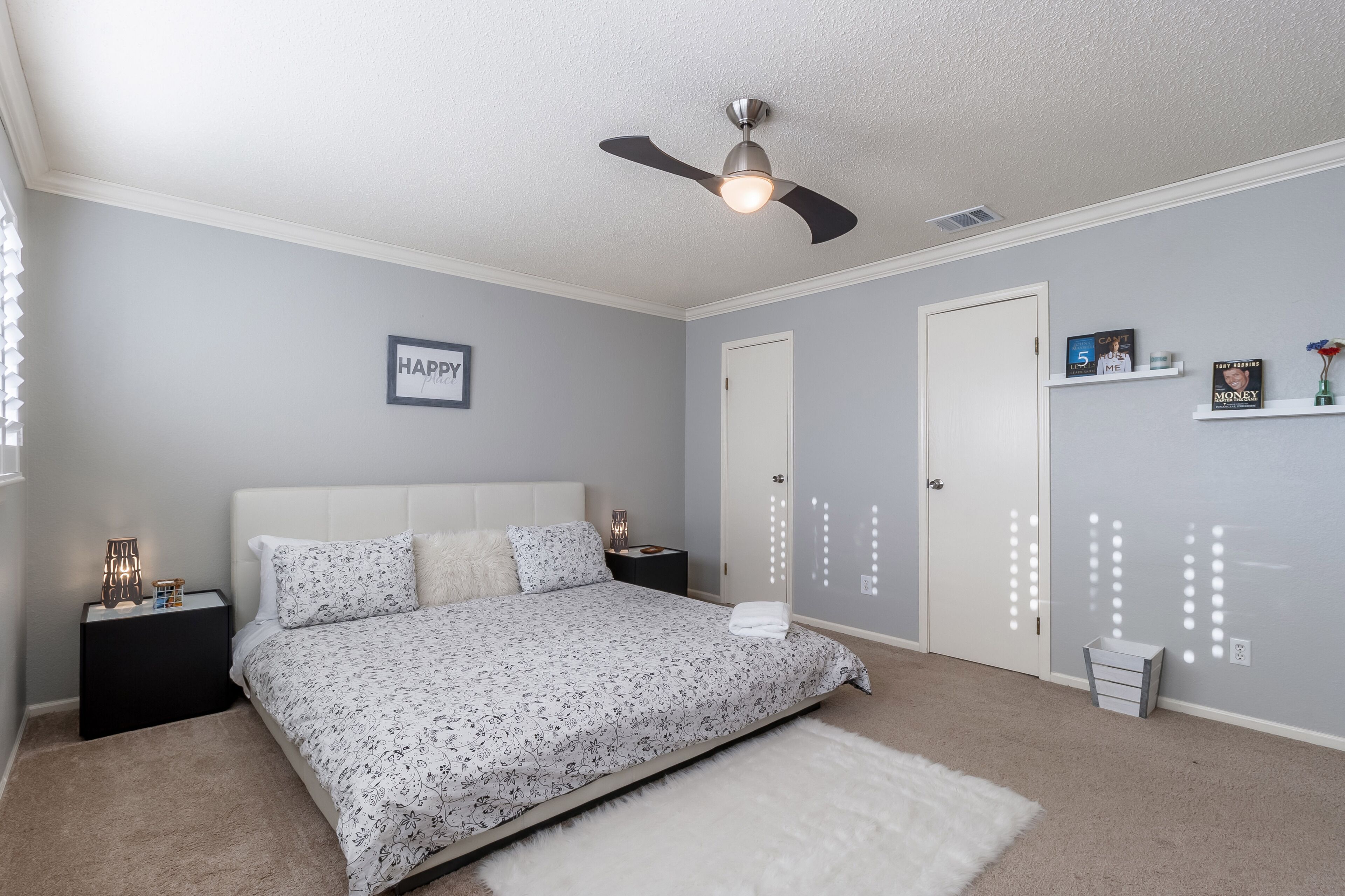 Quarto com cama, mesas de cabeceira, ventilador de teto, porta branca e detalhes decorativos nas paredes.