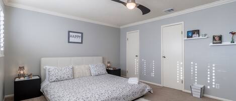 Quarto com cama, mesas de cabeceira, ventilador de teto, porta branca e detalhes decorativos nas paredes.