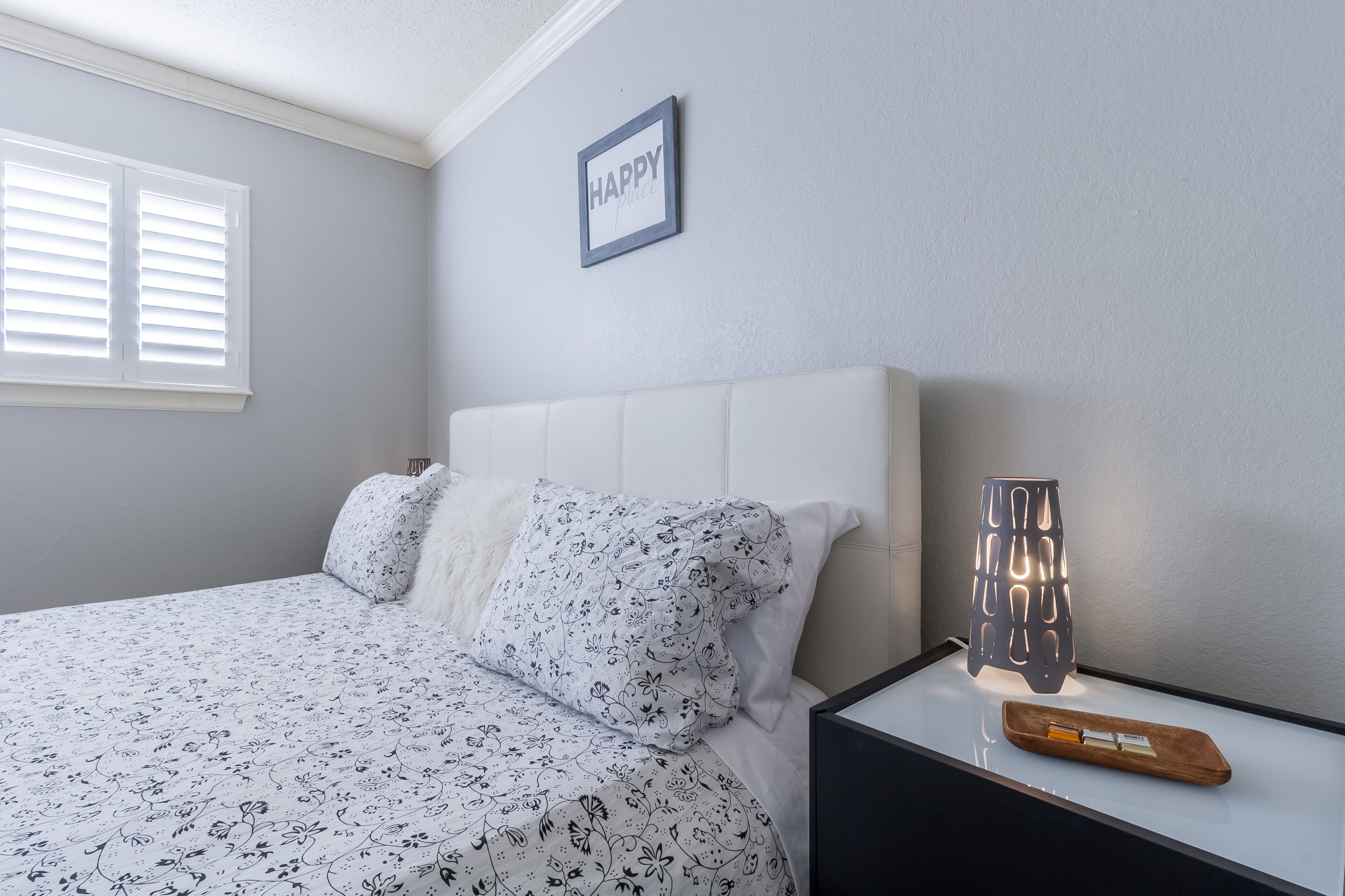 Een slaapkamer met een bed, kussens, een nachtkastje met een lamp, en een bordje met de tekst 'HAPPY' aan de muur.