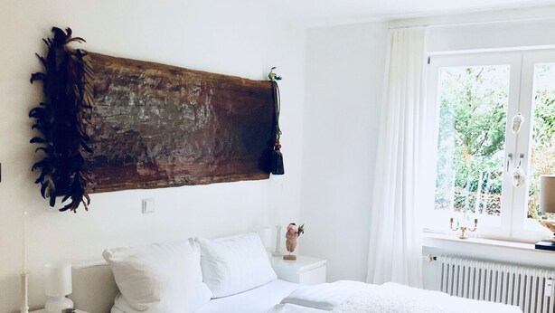 A bedroom with a large bed, white bedding, a wooden headboard, a nightstand with a lamp, and a window with a view of greenery. - very quiet apartment close to the sea and the center (Überlingen)