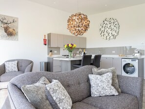 A modern living room with grey sofas, a dining area, a kitchen with a built-in washing machine, and decorative wall art.