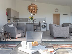 A modern living room with grey sofas, a dining table with a breakfast setting, a kitchen area, and a large decorative light fixture.
