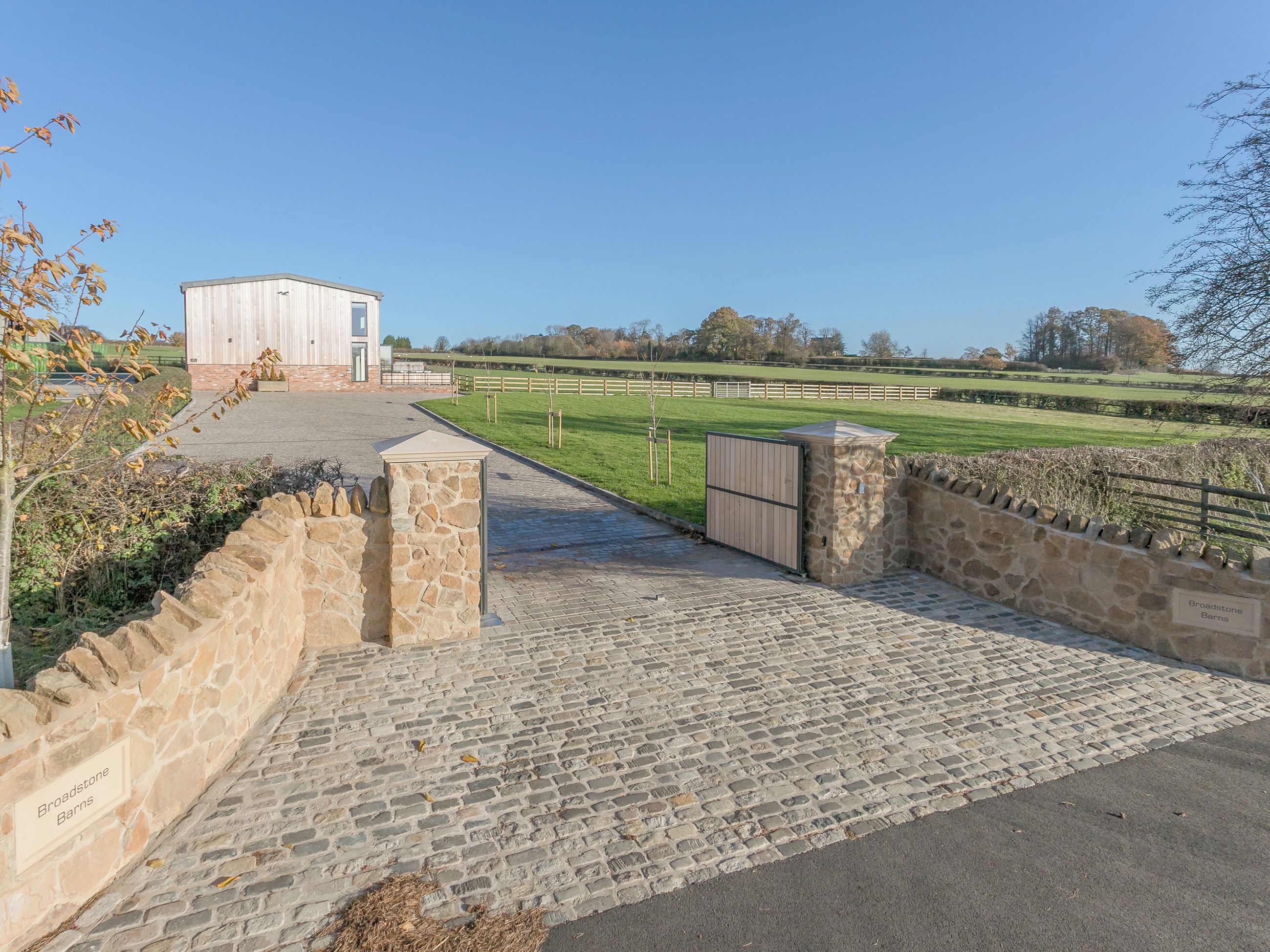 A paved entrance with stone walls, a wooden gate, and a building labeled 'Broadstone Barns' in the background.