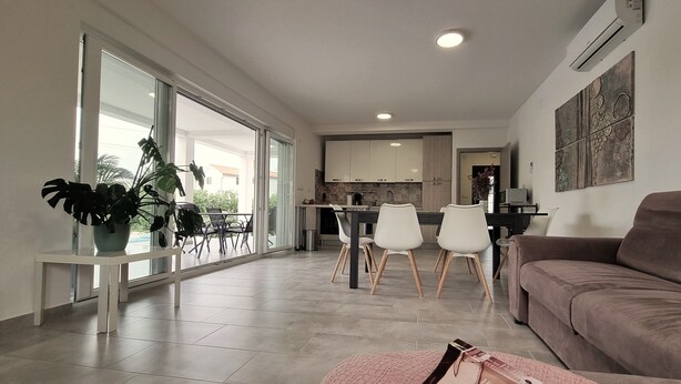 A modern living room with a large window, a dining area with chairs, a kitchen in the background, and a brown sofa. - Aria, villa in Filipana, close to numerous beaches and activities (Marcana)