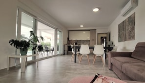 Una sala de estar moderna con un ventanal grande, un comedor con sillas, una cocina al fondo y un sofá marrón.